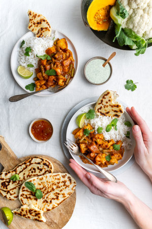 Top down down view of a vegetarian, vegan Indian Balti curry with cauliflower and pumpkin served with raita, naan bread and mango chutney and basmati rice on white background, female hands holding one plateの写真素材