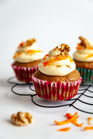 three carrot cake muffins with icing and carrot shaving on cooling rack on a white backgroundの写真素材