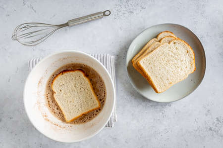 Making process of French toast, one slice of bread soaking in bowl with plate of bread and whisk next to it on white gray background, minimal flat lay with copyspaceの写真素材