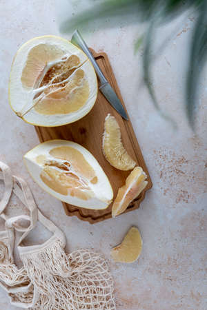 Bird's eye view of sliced yellow pomelo with wedges on a wooden cutting board with mesh bag and bamboo on neutral background with copy spaceの写真素材