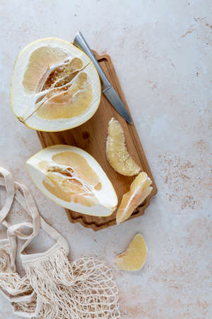 Bird's eye view of sliced yellow pomelo with wedges on a wooden cutting board with mesh bag on neutral background with copy spaceの写真素材