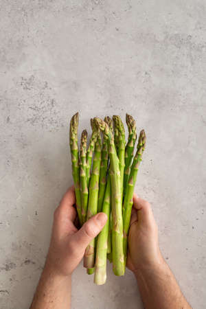 A man holding a bunch of fresh raw green asparagus on gray background with copy space above, verticalの写真素材
