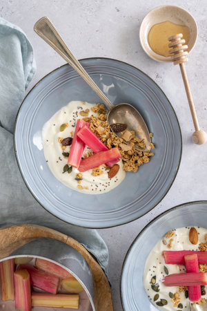 Flat lay of yoghurt and poached rhubarb with granola in two blue bowls with honey, healthy breakfast, vertical on a gray backgroundの写真素材