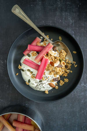 Flat lay of yoghurt and poached rhubarb with granola in a black bowl and a pan with rhubarb on black background, with copy space, verticalの写真素材
