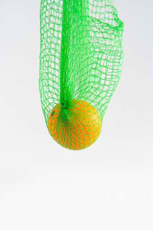 A single orange hanging in a green net bag on white background with copy space, horizontal, food shoppingの写真素材