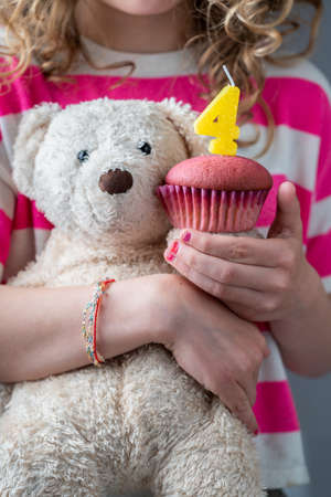 Celebrating a girl's fourth birthday, a girl in a pink and white shirt holding a white teddy bear and a pink cupcake with a candle with the number four, close-upの写真素材