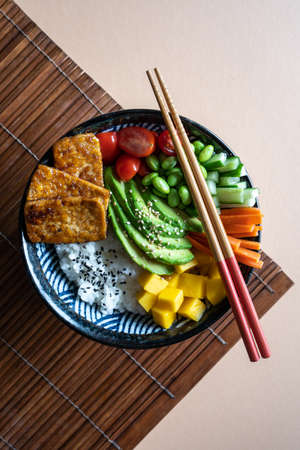 Flatlay of a homemade vegan poke bowl with tofu, mango, avocado, carrot, edamame, tomato, cucumber on a almond and bamboo mat background , verticalの写真素材