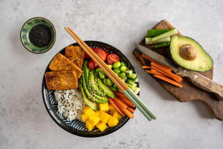 Top down view of a homemade vegan poke bowl with tofu, mango, avocado, carrot, edamame, tomato,...