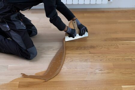 Carpenter master with gloves grouting wooden parquet, phase after sanding. Varnishing of oak parquet floor, man workers with hand and spatulaの写真素材