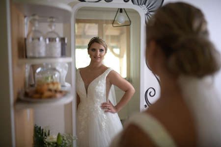 A elegant and beautiful bride at home. Beauty portrait of bride wearing fashion wedding dress with luxury delight make-up and hairstyle in mirrorの写真素材