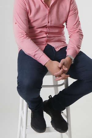 A young attractive guy in a bright red shirt and jeans sits on a bar stool on a white backgroundの写真素材