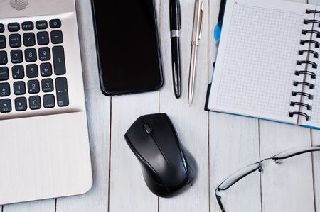 Laptop with mouse, smartphone, notebook, pens and glasses on white wooden table. Business concept