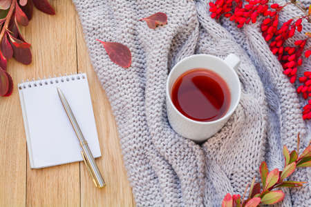 Autumn flatlay on a wooden background with a cup of tea and branches of barberry. Notepad with pen...