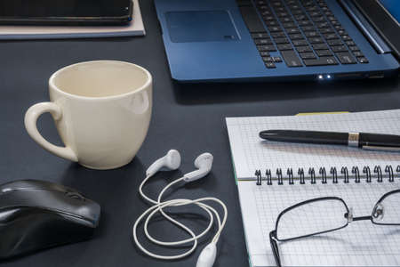 Laptop with mouse, coffee cup, notebook with pen and other accessories on the desktop with a dark...