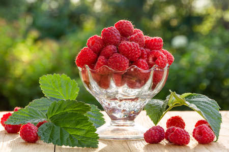 Fresh ripe raspberry berries in a transparent glass bowl on a wooden table in the garden