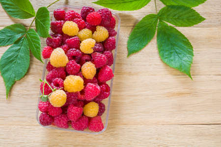 Fresh ripe red and yellow raspberry berries in a transparent plastic container on a wooden table