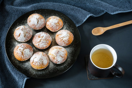 Freshly baked appetizing cupcakes sprinkled with powdered sugar on a black plate and a cup of tea on a table with a dark surfaceの写真素材