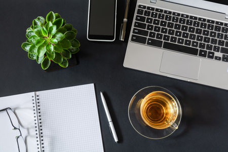 Workplace with laptop, notebook with pen, cup of tea and smartphone on dark desktop. Flat lay, top view office deskの写真素材