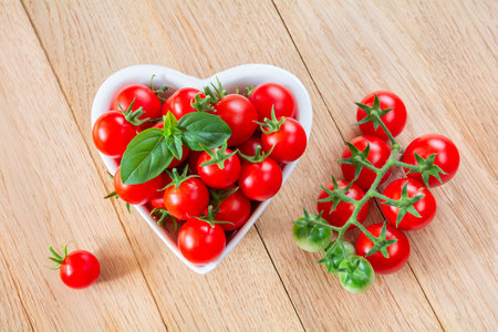 Red ripe cherry tomatoes in a white heart-shaped bowl on a wooden backgroundの写真素材