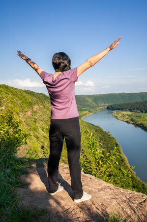 A young happy woman is standing on a stone on a high river bank with her arms raisedの写真素材