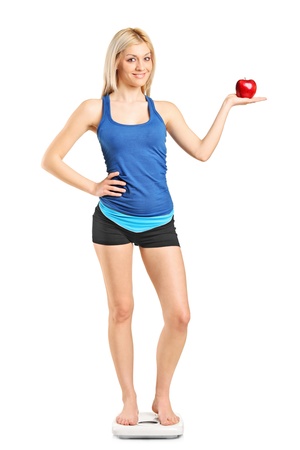 Full length portrait of a smiling woman holding a red apple and standing on a weight scale isolated on white backgroundの写真素材