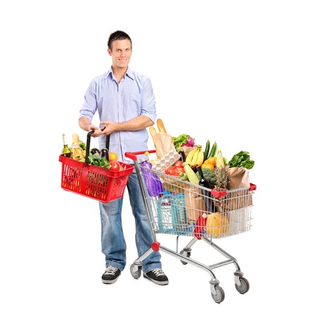 Full length portrait of a young man holding a basket full with products and shopping cart isolated on whte backgroundの写真素材