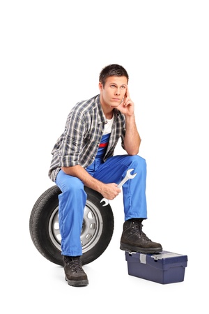 A thoughtfull mechanic sitting on a spare tire and holding a wrench isolated on white backgroundの写真素材