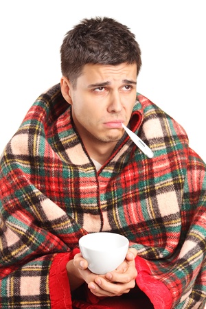 Ill man covered with blanket holding a tea cup and a thermometer in his mouth isolated against white backgroundの写真素材