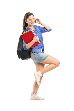 Full length portrait of a smiling school girl holding books and talking on a phone isolated on white backgroundの写真素材