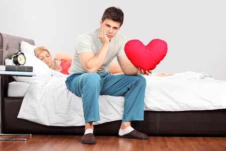 Young man in thoughts holding a heart shaped pillow while his girlfriend fall asleepの写真素材