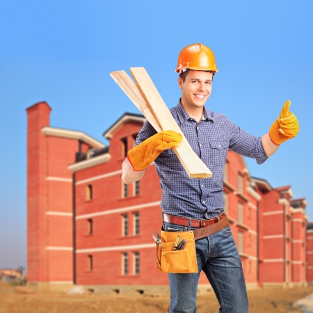 A smiling carpenter holding sills and giving thumb up with a newly built apartment block in the backgroundの写真素材