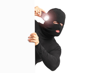 A thief with robbery mask holding a flashlight behind a white panel isolated on white backgroundの写真素材