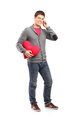 Full length portrait of a young male holding a red heart shaped pillow and talking on a phone isolated on white backgroundの写真素材