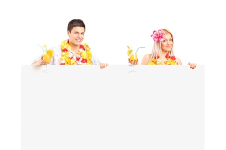 Young couple dressed in a hawaiian costume holding a cocktails and posing behind a white panelの写真素材