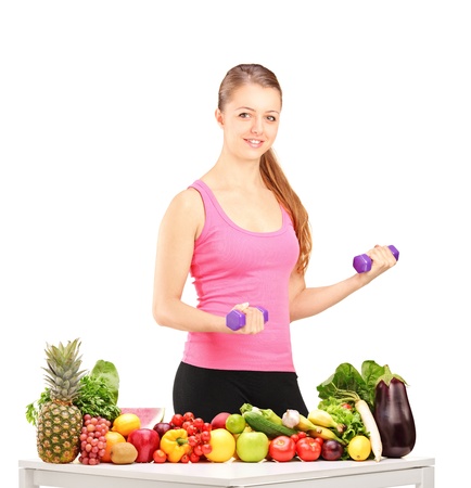 Smiling woman holding weights and table full of fruits and vegetables isolated on white backgroundの写真素材