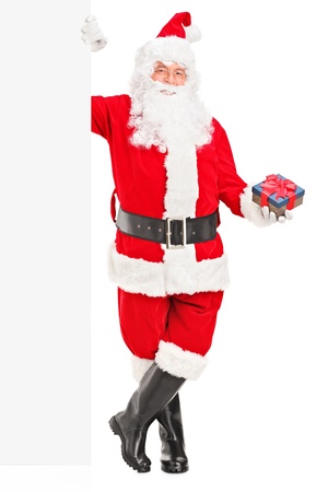 Full length portrait of a happy Santa claus standing next to a blank billboard and holding a gift isolated on whiteの写真素材
