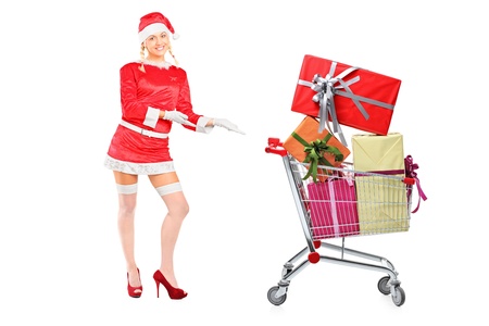 Full length portrait of a sexy woman wearing santa costume and showing a shopping cart full of presents isolated on white background の写真素材