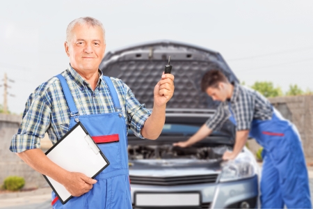 Mature mechanic in uniform holding a car key and another mechanic performing a car check in the backgroundの写真素材