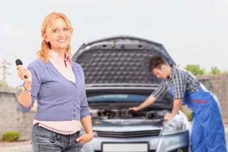 Smiling careless female holding a key while in the background mechanic is checking her carの写真素材