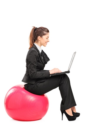 A busy woman sitting on a pilates ball and working on a notebook computer isolated against white backgroundの写真素材