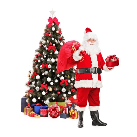 Full length portrait of a Santa Claus holding a bag and standing next to a Christmas tree isolated on white backgroundの写真素材