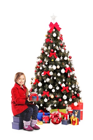 Happy little girl sitting next to a christmas tree and holding present isolated on white backgroundの写真素材