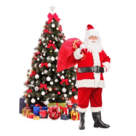 Full length portrait of Santa Claus carrying a bag full of gifts on his back, christmas tree in the backgroundの写真素材