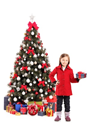 Happy little girl standing next to a christmas tree and holding present isolated on white backgroundの写真素材