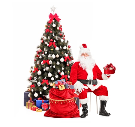 Santa Claus with a gift sitting next to a bag full of presents, christmas tree in the backgroundの写真素材