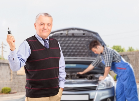 Smiling mature gentleman holding a car key while in the background mechanic is checking his carの写真素材