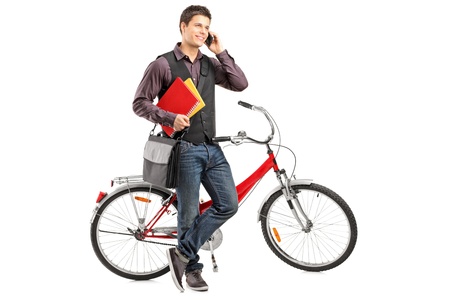Full length portrait of a smiling student holding books and talking on a phone next to a bike isolated on white backgroundの写真素材