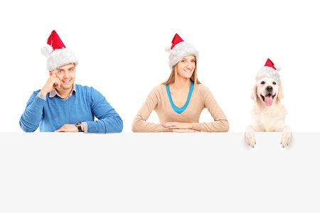 Smiling couple and dog with santa claus hats posing behind a blank panelの写真素材