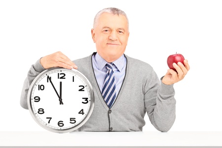 Smiling gentleman sitting and holding a wall clock and a red apple isolated on white backgroundの写真素材