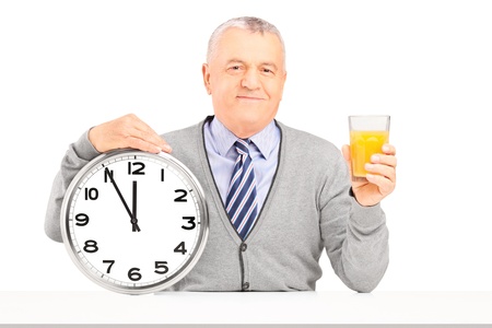Smiling gentleman sitting and holding a wall clock and glass of orange juice isolated on white...